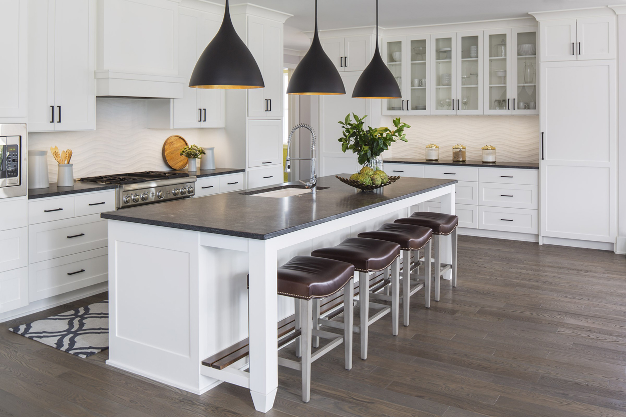 White kitchen with dark hickory wood floors. Kitchen with black pendant lights over white kitchen island with black marble countertop 