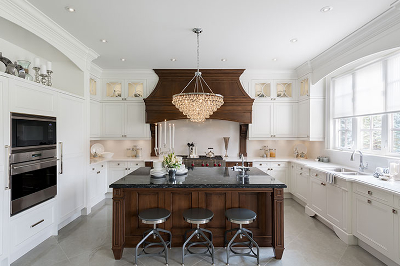 White kitchen with iron swivel bar stools and wooden kitchen island. Kitchen with crystal chandelier over wooden kitchen island with black granite countertop