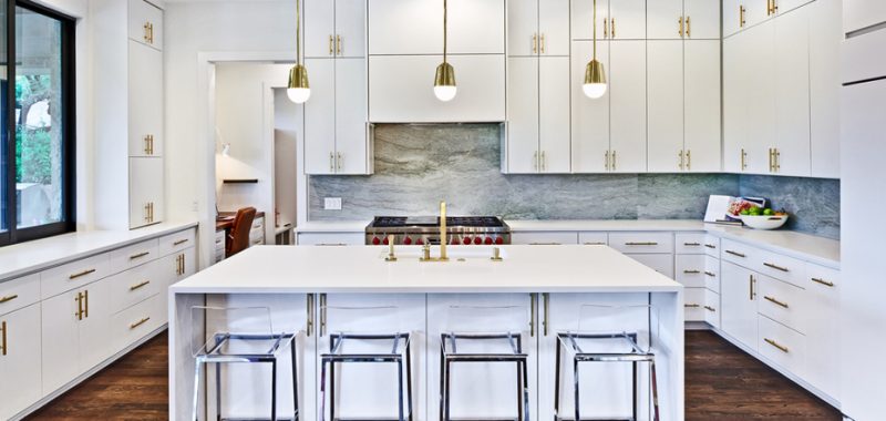White kitchen with marble tile backsplash and glass bar stools. Kitchen with mini gold pendant lights over white kitchen island with laminate countertops
