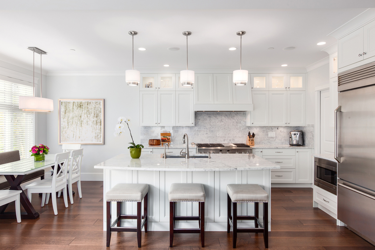 White kitchen design with bar stools, dining area and dark hardwood flooring. Kitchen with small drum pendant light over white kitchen island with white marble countertop